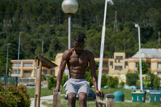 Determined African American male athlete with naked torso balancing on parallel bars and doing abs exercises during intense workout on sports ground in summer