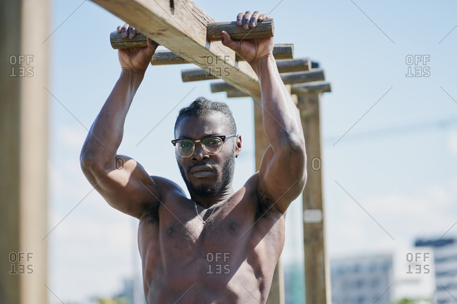 Low angle of serious strong shirtless African American male in eyeglasses doing calisthenics exercise on monkey bars during intense bodyweight outdoor workout