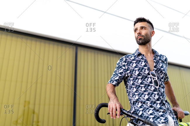 From below serious handsome male in stylish clothing standing with modern bike near modern wall in city while looking away