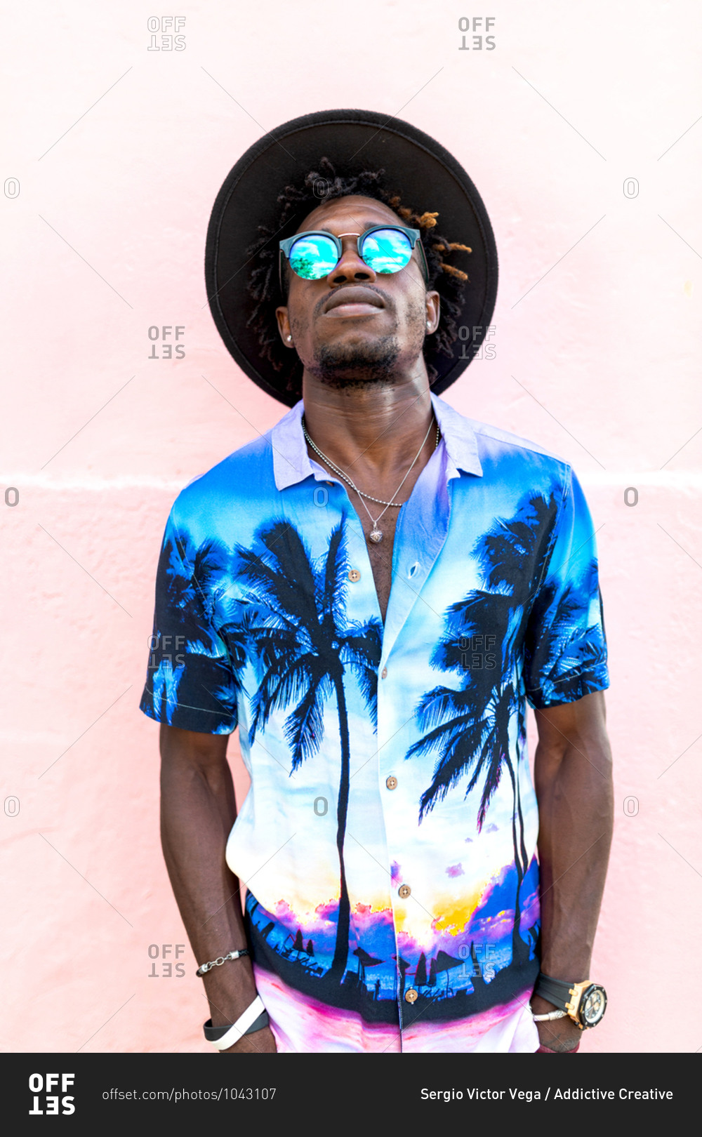 Confident African American male wearing trendy shirt with palm trees and stylish sunglasses standing on street near building with pink wall