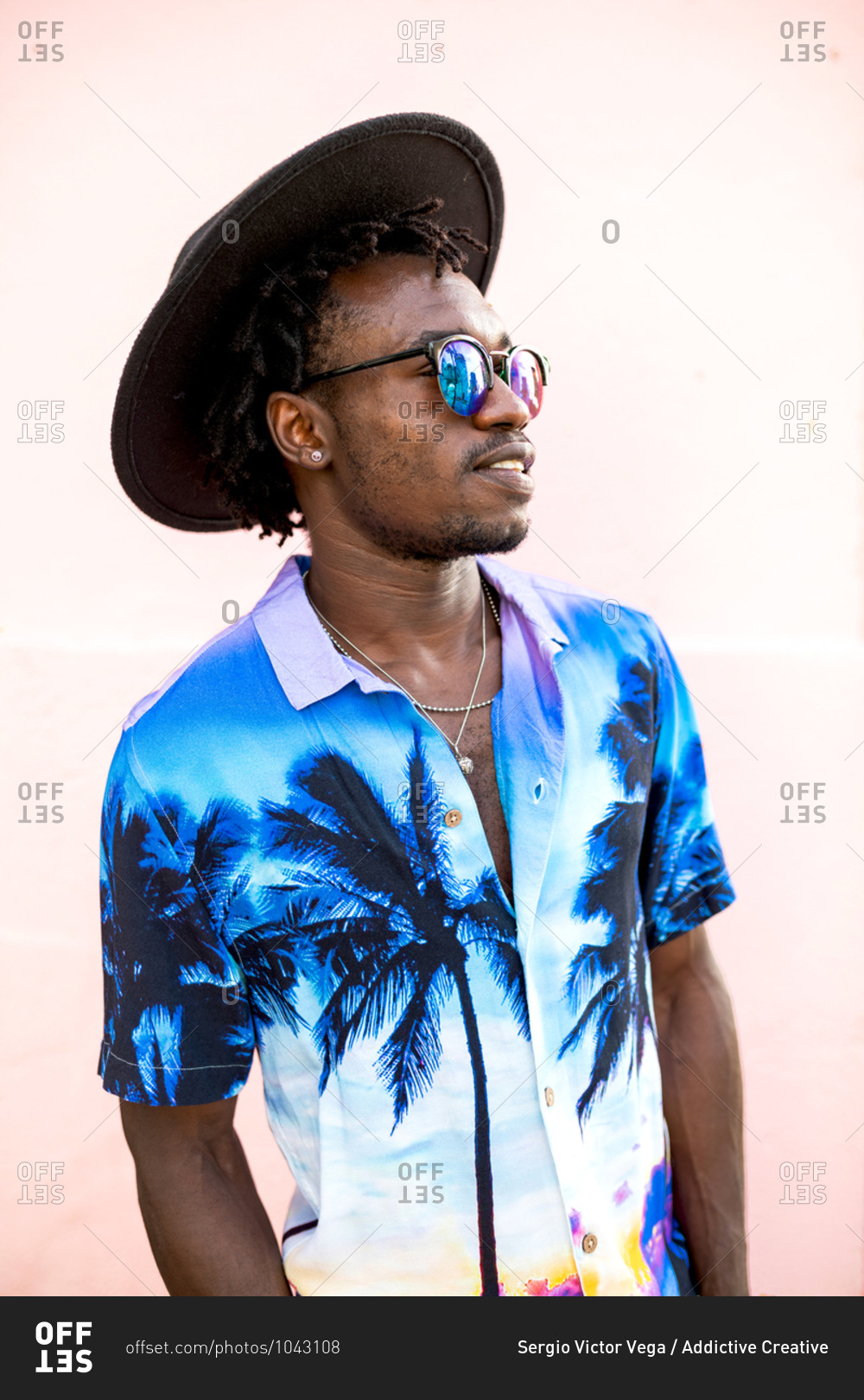 Confident African American male wearing trendy shirt with palm trees, hat and stylish sunglasses standing on street near building with pink wall