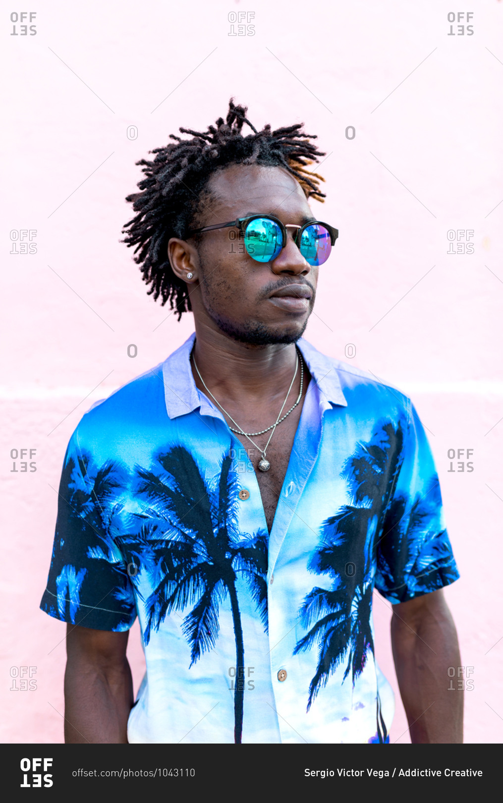 Confident African American male wearing trendy shirt with palm trees and stylish sunglasses standing on street near building with pink wall