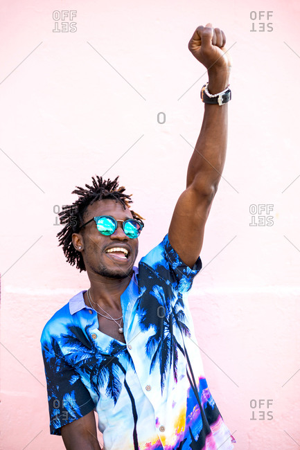Stylish African American male in trendy outfit and sunglasses standing in city with raised arm and celebrating success while smiling brightly