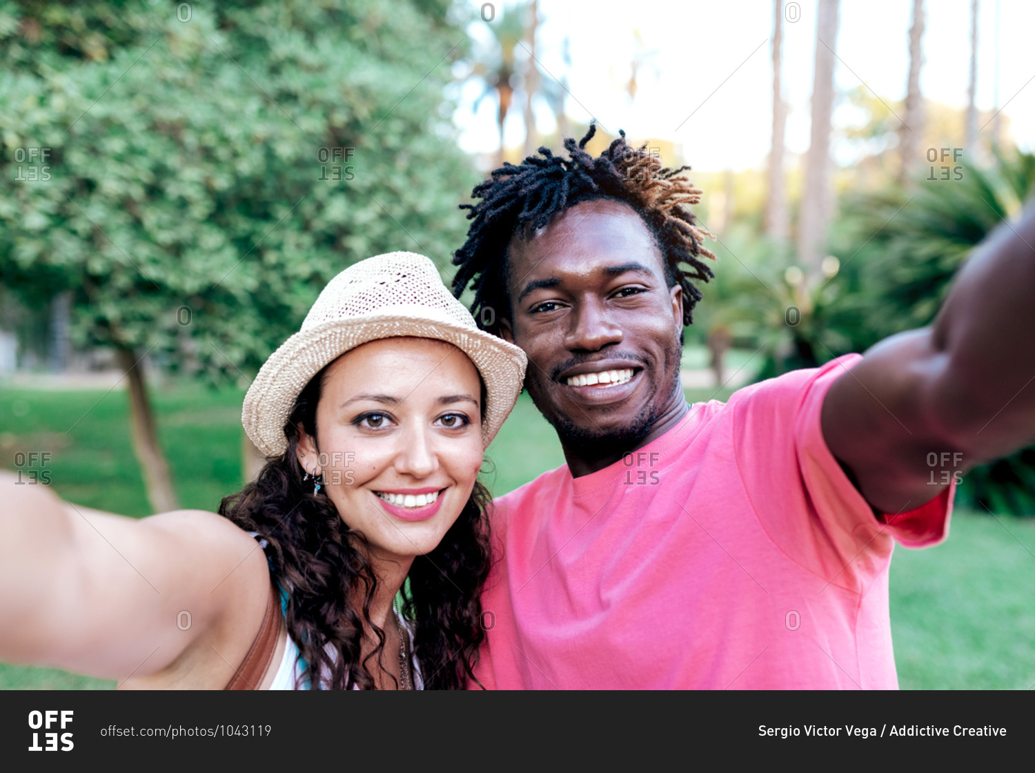 Cheerful multiracial traveling couple hugging and taking selfie on smartphone while standing in green tropical park during summer vacation