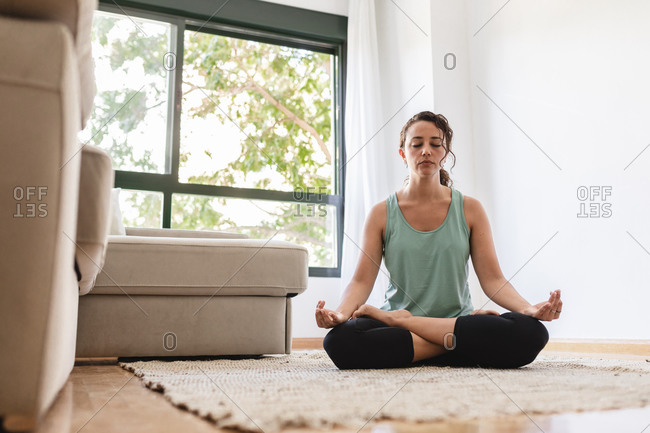 Peaceful female in sportswear practicing yoga in lotus pose while sitting on carpet in living room