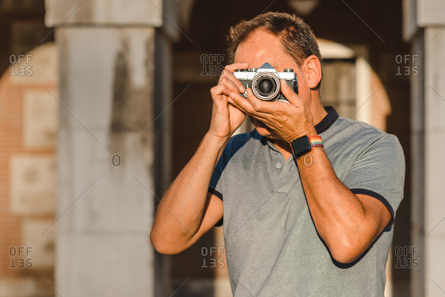 Unrecognizable male photographer covering face with retro photo camera and taking photo on sunny day on street