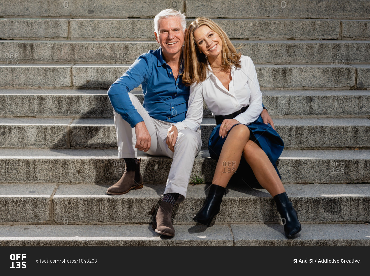 Mature couple in love sitting on stone steps in city and leaning on each other while looking at camera and enjoying stroll at weekend