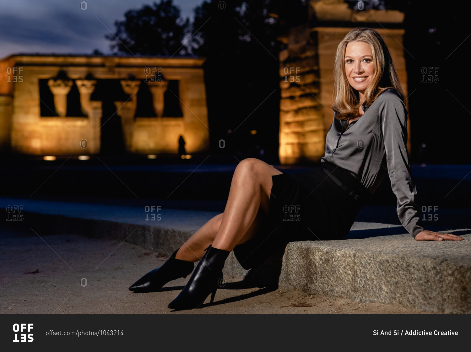Side view full body of happy confident middle aged female in stylish outfit and trendy footwear sitting on stone border in night city