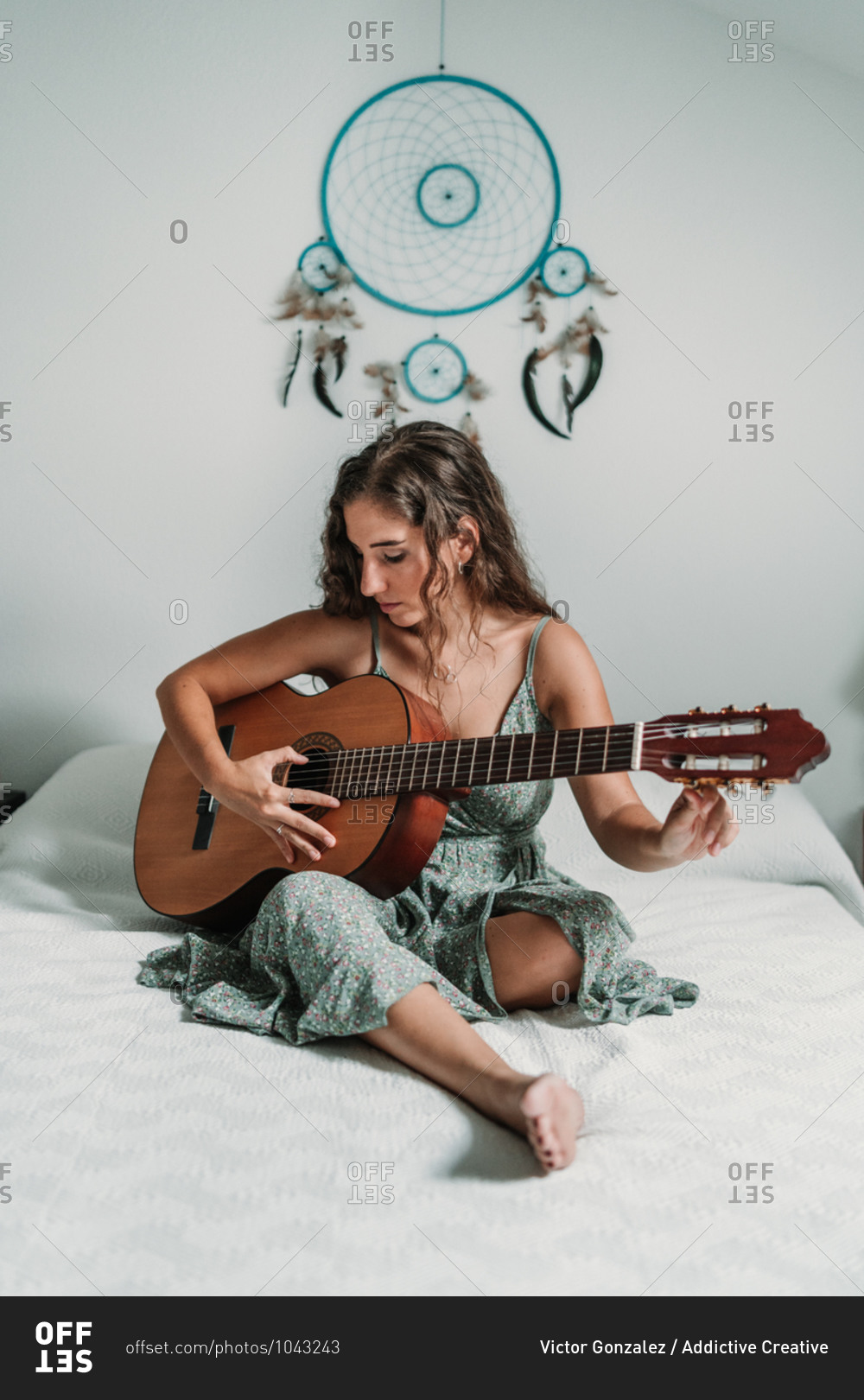 Female guitarist sitting on cozy bed at home and playing acoustic musical instrument with closed eyes