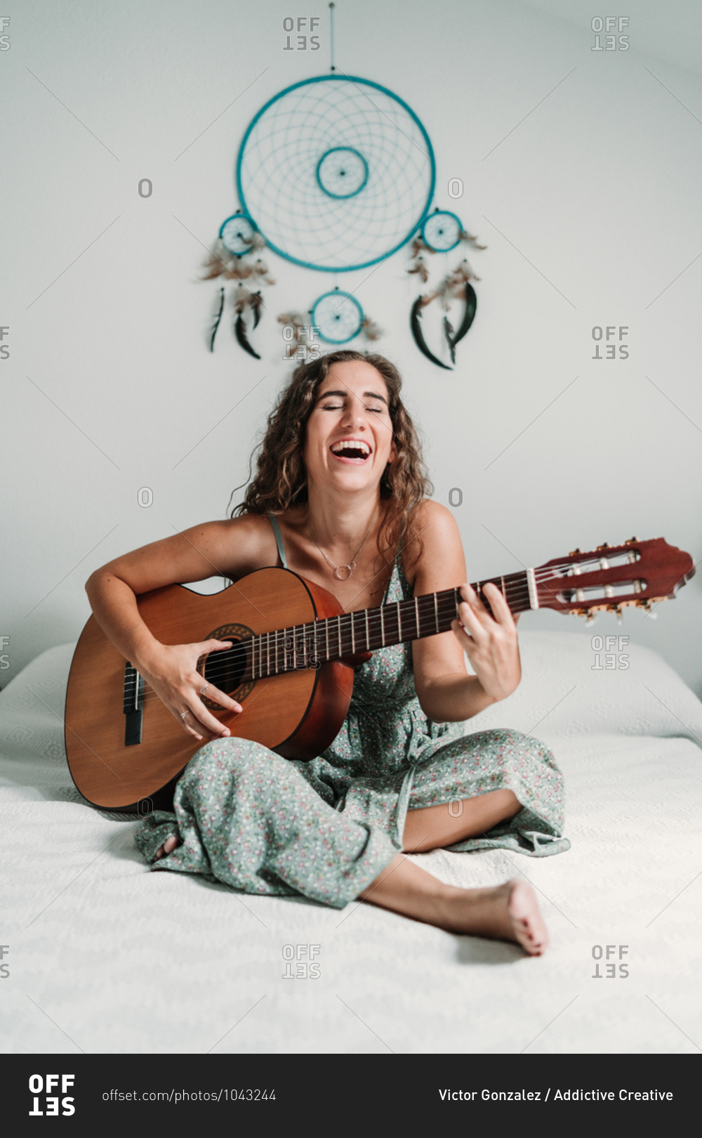 Cheerful ethnic female guitarist sitting on cozy bed at home and playing acoustic musical instrument with closed eyes
