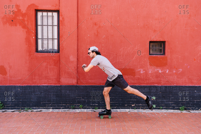 Side view of active stylish male in roller skates skating along street near red building while entertaining at weekend in summer