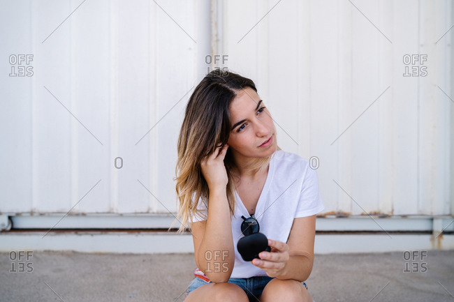 Modern millennial female in casual outfit adjusting wireless earphones while sitting against metal wall on city street