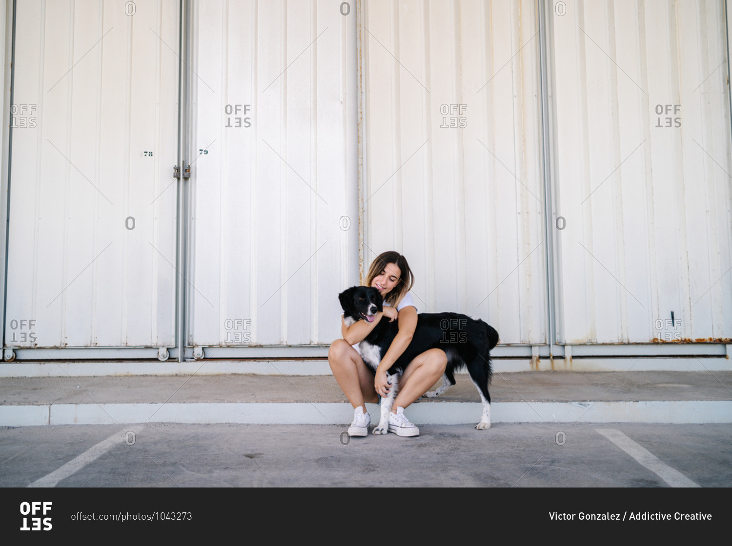 Full body side view of young female in casual outfit sitting with big dog near metal wall of urban building