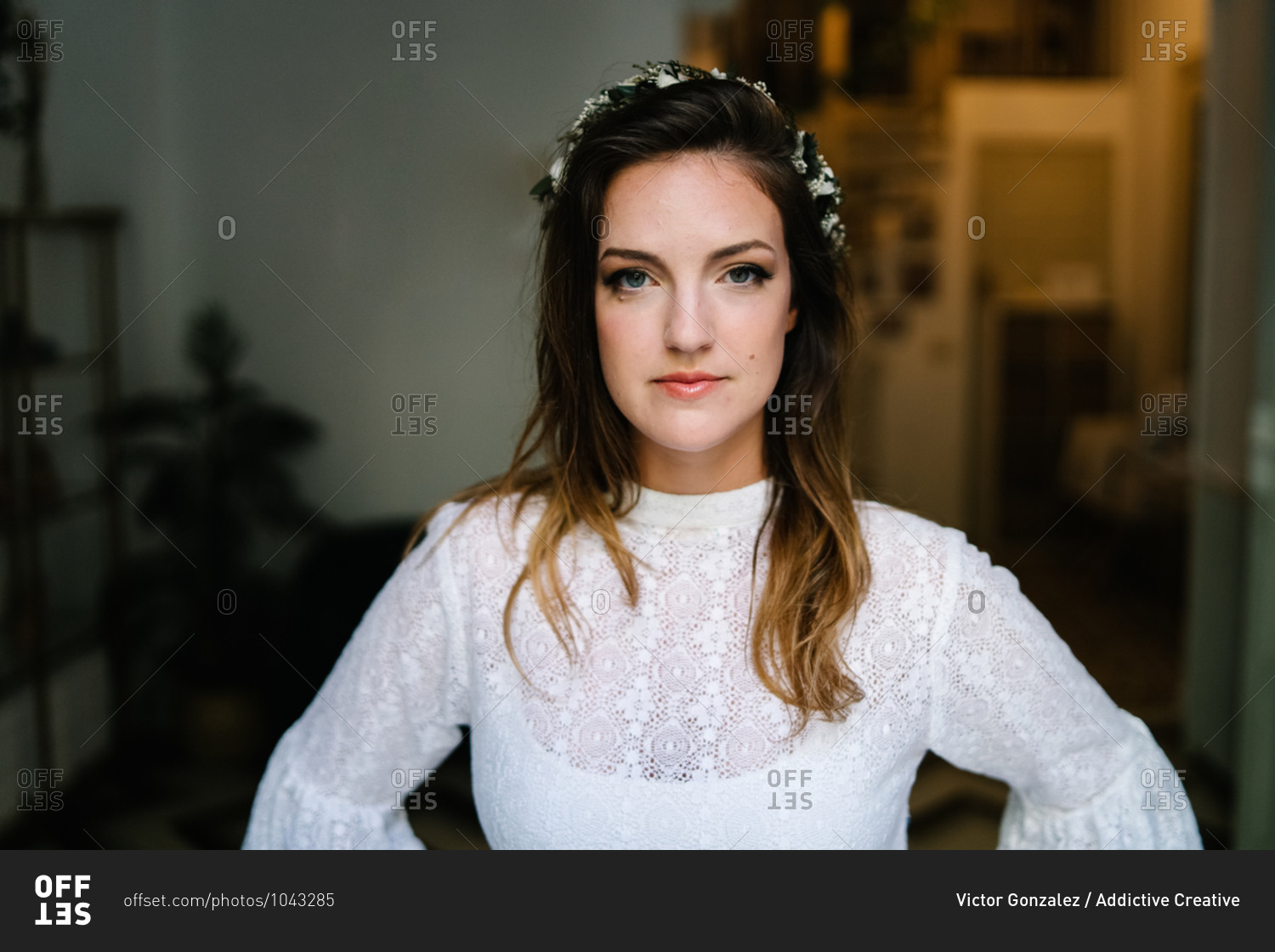 Charming female in elegant white dress and with floral wreath in hair standing in flat and looking at camera