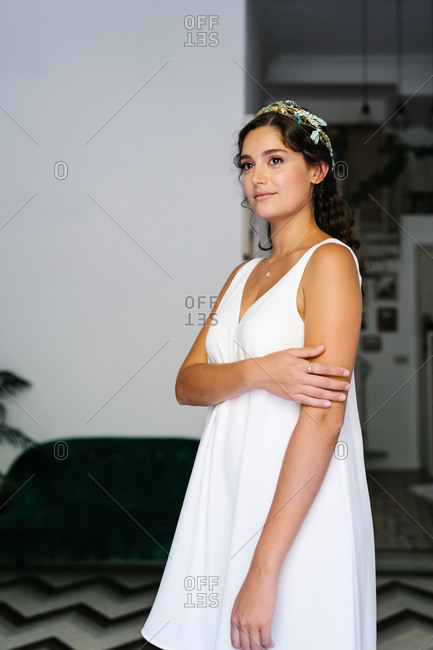 Tender female wearing delicate white dress standing in modern apartment and looking away