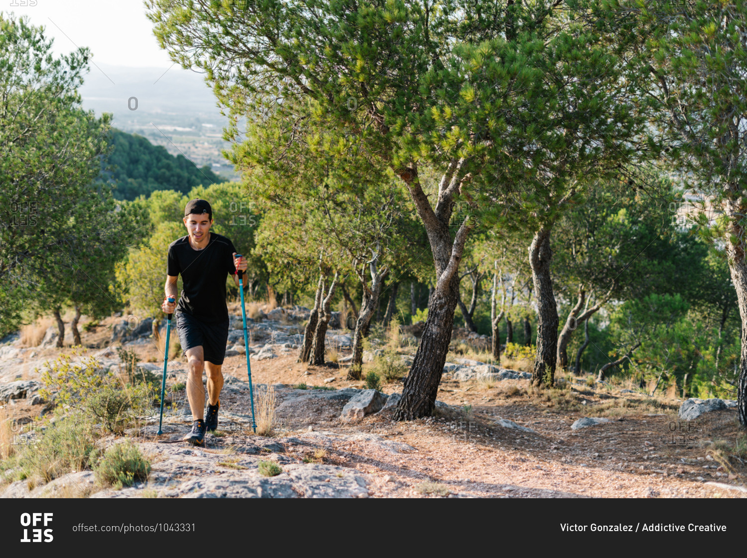Positive young athletic man in activewear hiking with trekking poles on stony trail in green forested mountainous terrain