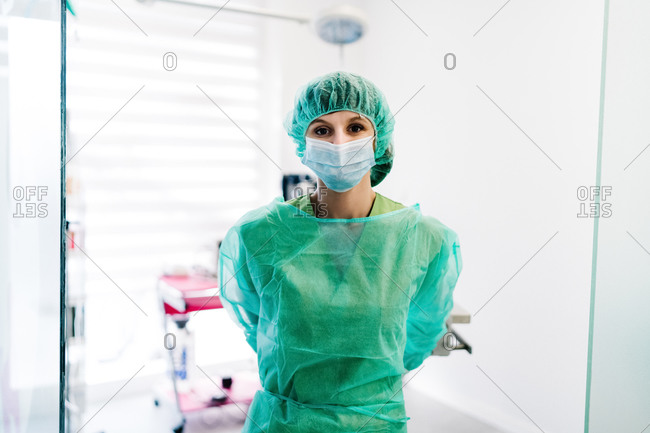 Serious female vet surgeon in mask and uniform preparing for operation while standing in operating theater of veterinary hospital and looking at camera