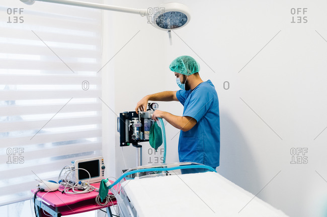 Side view of male vet doctor in uniform and mask preparing anesthesia machine for surgery in bright operating theater in veterinary clinic
