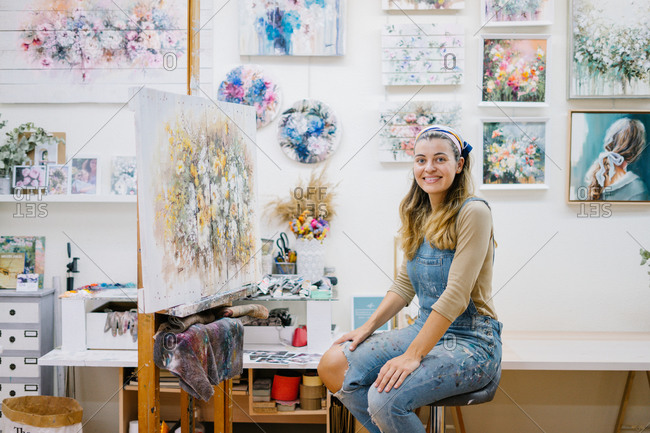 Side view of cheerful female artist in denim overall looking at camera while sitting near easel and painting picture with paintbrush in creative atelier