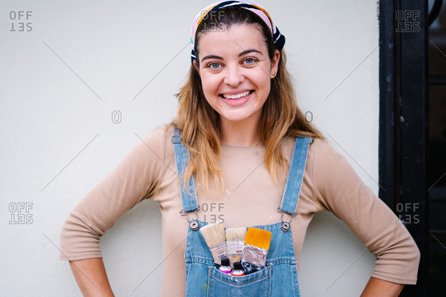 Cheerful young female artist with paintbrushes and paint tubes in pocket of denim overall looking at camera while standing against white wall in workshop