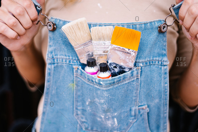 Crop anonymous female artist in denim overall with various paintbrushes and colorful paint tubes in pocket