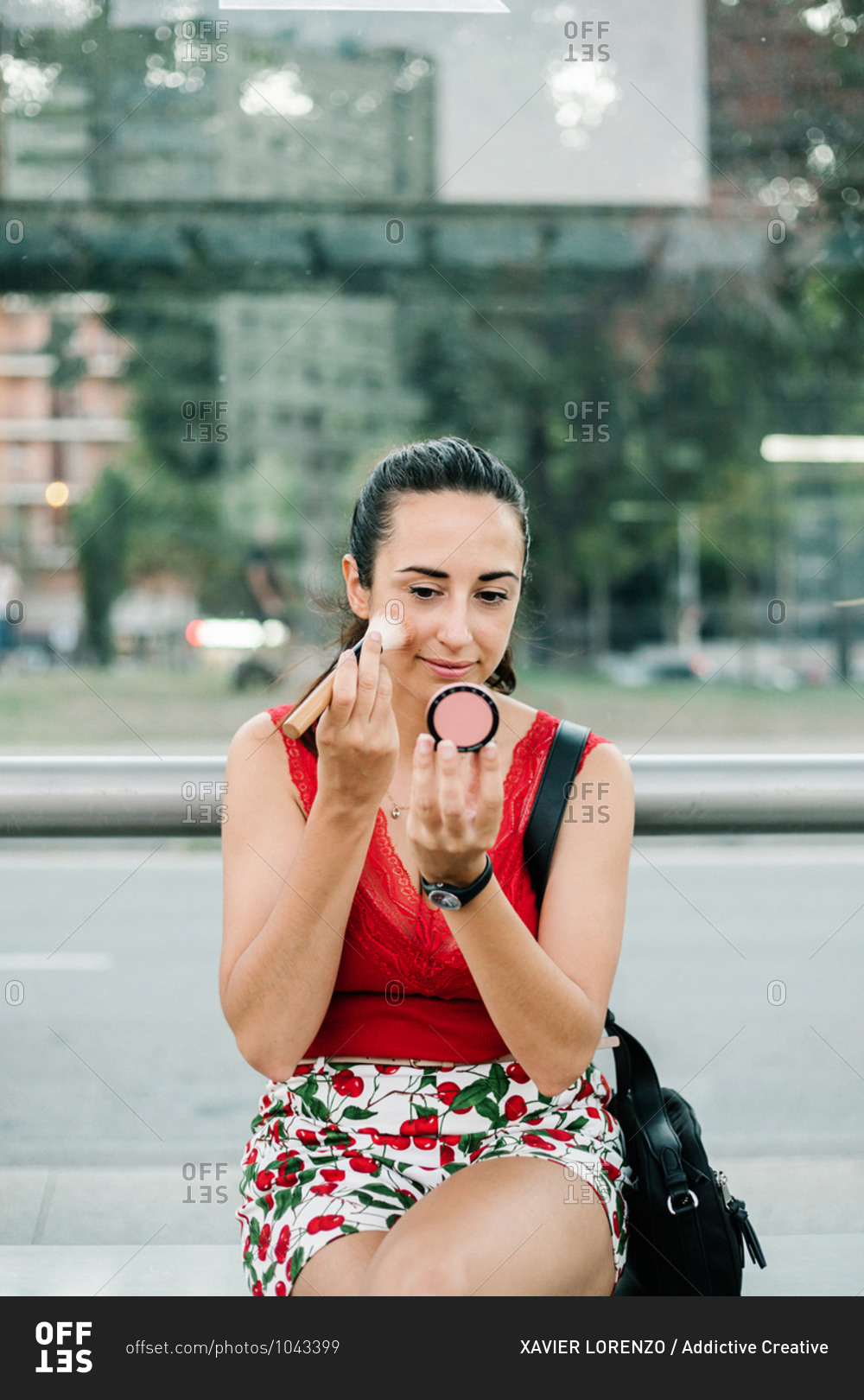 Modern brunette in red blouse looking at reflection on blush mirror while standing on city street and applying blush with brush