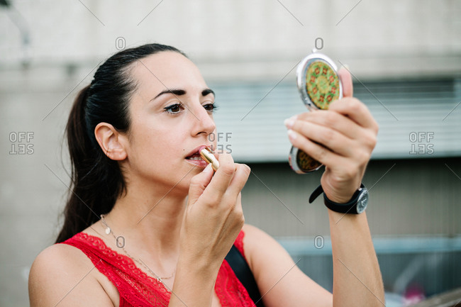 Modern brunette in red blouse looking at reflection on mirror while standing on city street and applying lipstick
