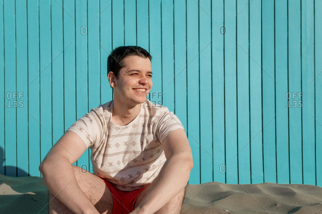 Young man wearing swimsuit pants smiling in sandy beach looking at sun