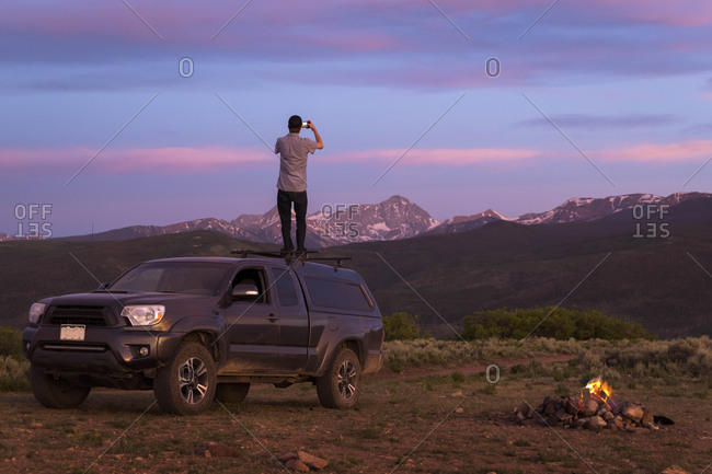 Rear view of man photographing while standing on off-road car during sunset
