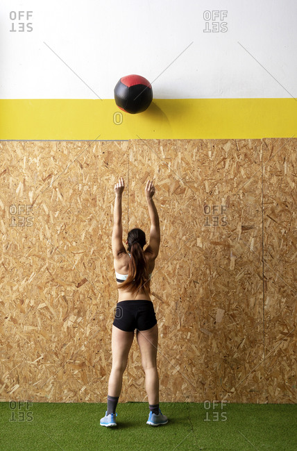 Rear view of beautiful young athlete with ponytail throwing up heavy leather ball to cork wall at gym