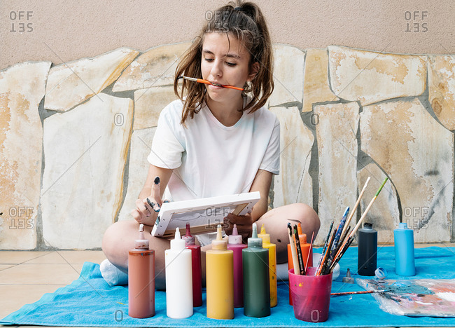 Caucasian girl biting a painting brush while painting with her finger, sitting on her terrace