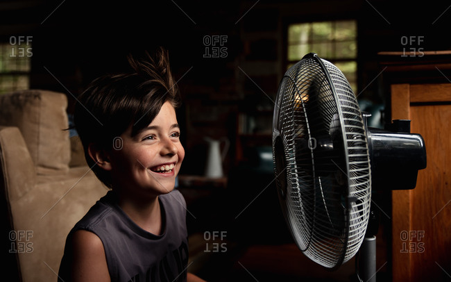 Young boy smiling as he is cooling off in front of a fan in dark room.