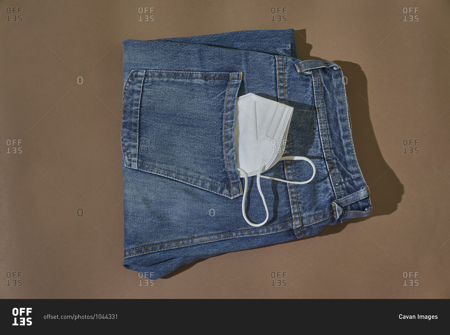 From above of folded jeans with white protective mask in pocket placed on brown background.