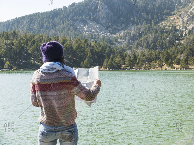hiker woman looking at map while standing in front of a lake in natural environment and thinking of where to go next