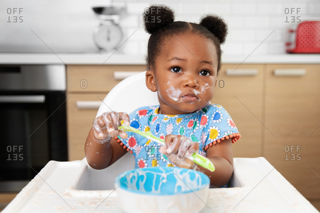 Messy baby girl in high chair eating yogurt with spoon