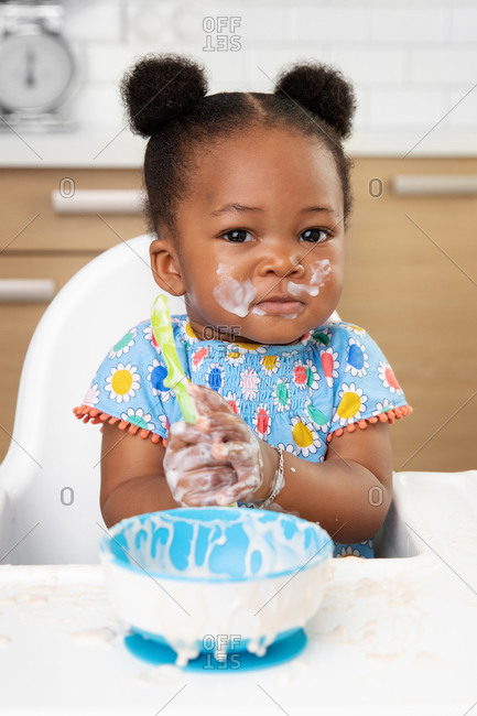 Funny baby girl with messy face eating yogurt with spoon in high chair