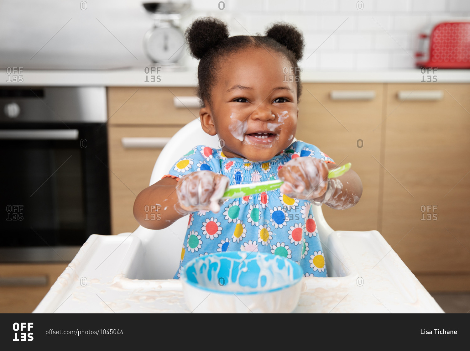 Laughing baby girl with messy face eating yogurt with spoon in high chair