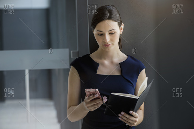 Confident businesswoman holding diary using smart phone while standing against wall