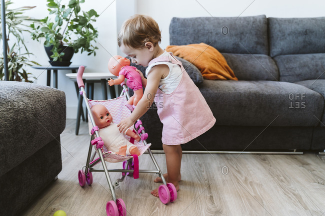 Cute baby girl putting toys in baby stroller while standing at home