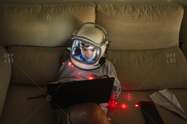 Boy wearing space helmet using laptop while sitting with illuminated lighting equipment on sofa at home