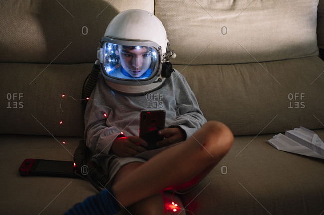 Boy wearing space helmet using smart phone while sitting with illuminated lighting equipment on sofa