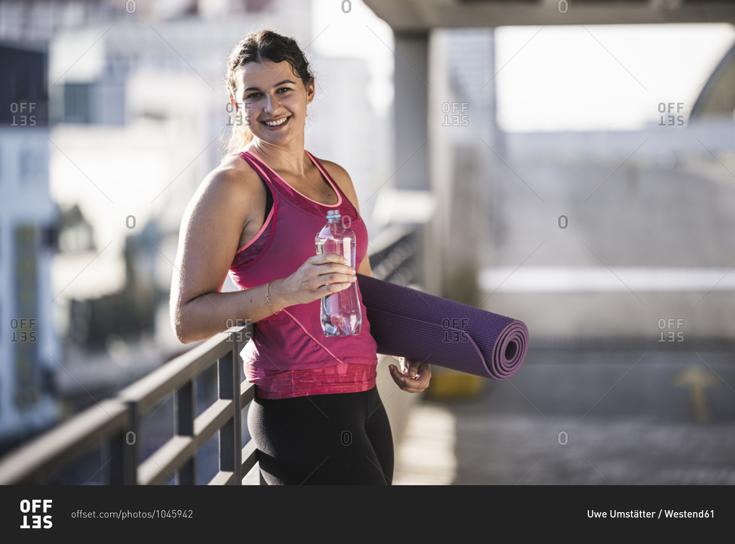 Young woman holding exercise mat and water bottle while standing by railing on terrace
