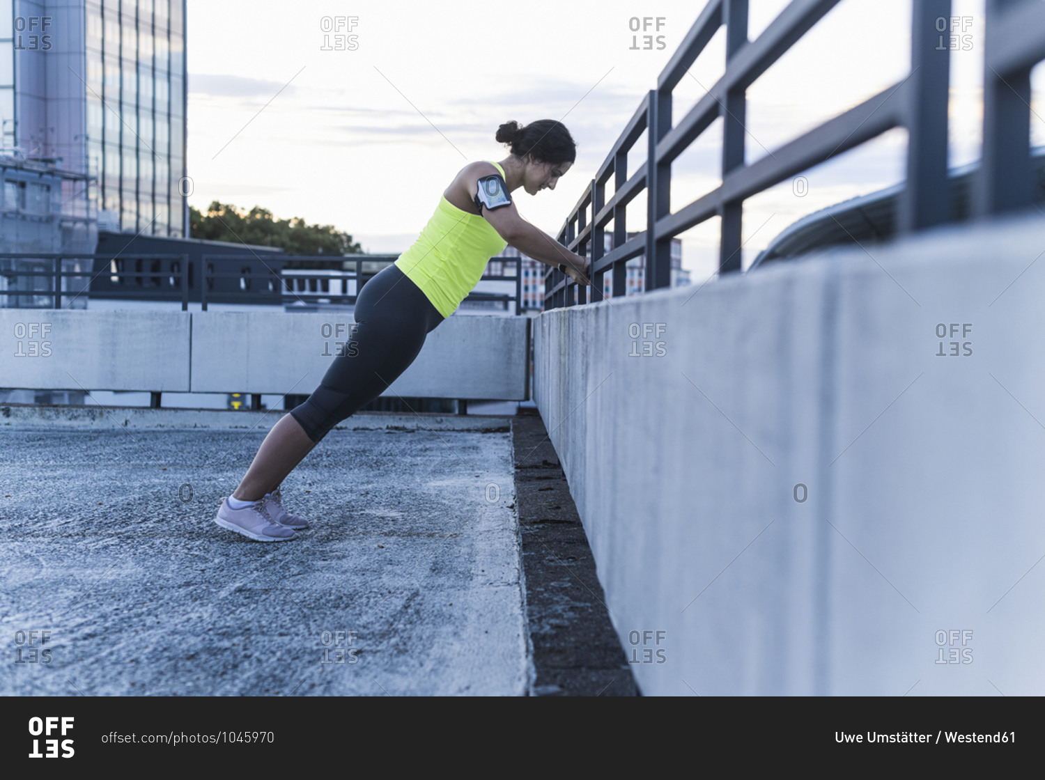 Young woman with arm band exercising on railing at sunset
