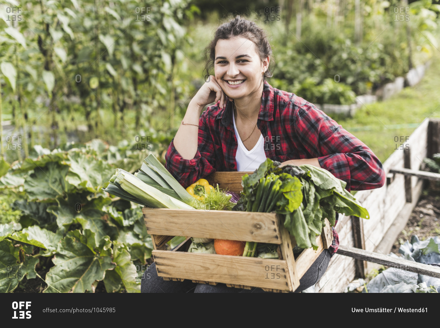 Smiling young woman sitting with vegetables in wooden crate at community garden