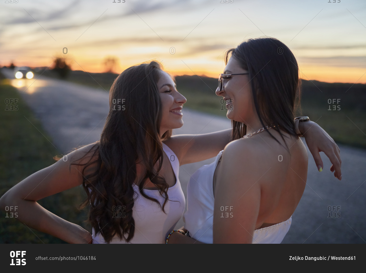 Smiling female friends looking at each other while standing on road against sky at sunset