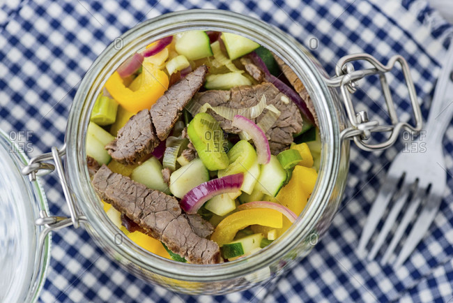 Close-up, mason jar with beef salad on blue and white checked cloth.