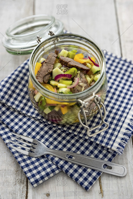 Beef salad, colorful vegetables, in a mason jar on a white wooden background and blue and white checked cloth, with a camping fork