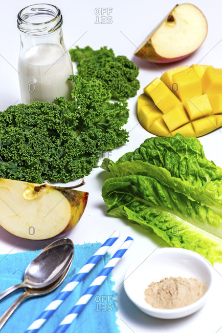 All ingredients spread out on a white background, milk bottle, kale, mango hedgehog, apple quarters, Romaine lettuce, two straws, two silver spoons, nutritional supplements in a white bowl