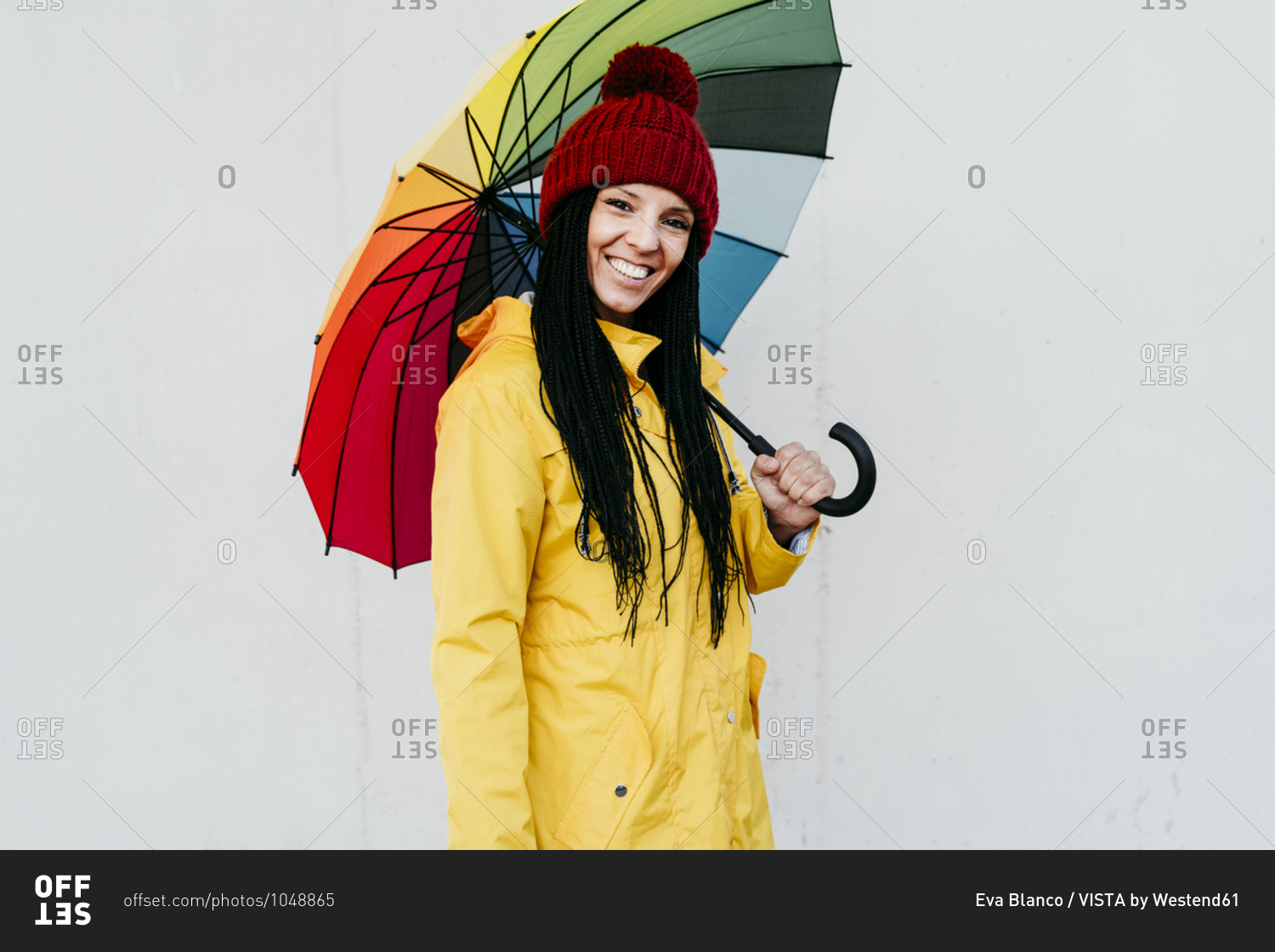 Smiling woman wearing raincoat holding colorful umbrella standing against wall