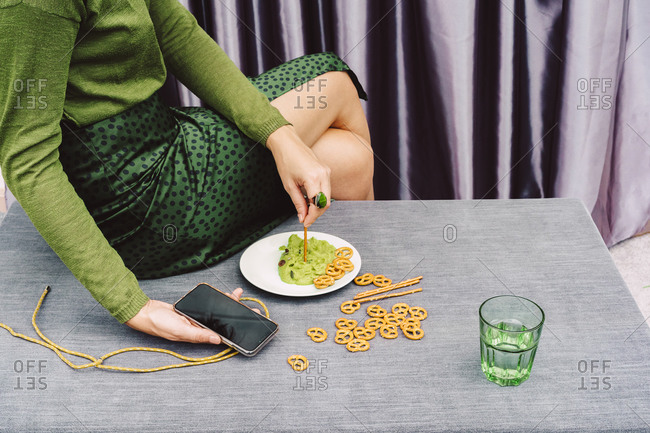 Woman holding smart phone eating guacamole and pretzel while sitting on table at home